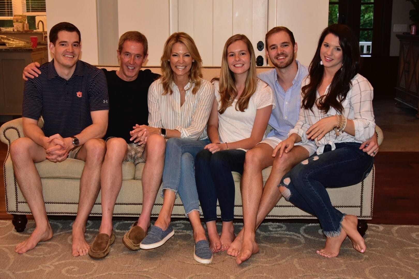 Family of six sitting together on a beige couch in a living room. The group includes parents and four adult children, all dressed casually and smiling. Kitchen area visible in the background