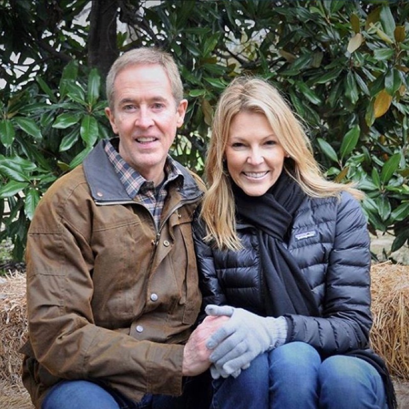 Couple sitting together outdoors against evergreen shrubs background. Man wearing brown jacket over plaid shirt, woman in black quilted jacket with blonde hair, both smiling at camera.