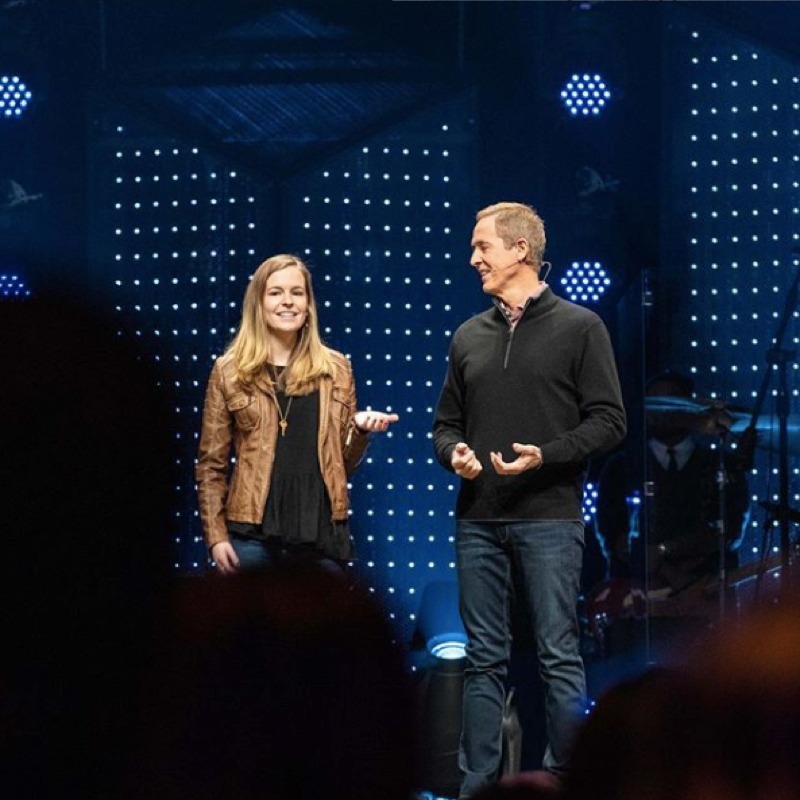 Two people on stage in what appears to be a church or auditorium setting with blue lighting and dotted backdrop. Young woman with long blonde hair wearing brown leather jacket over black top, man in dark quarter-zip pullover and jeans, both appearing to be speaking or presenting.