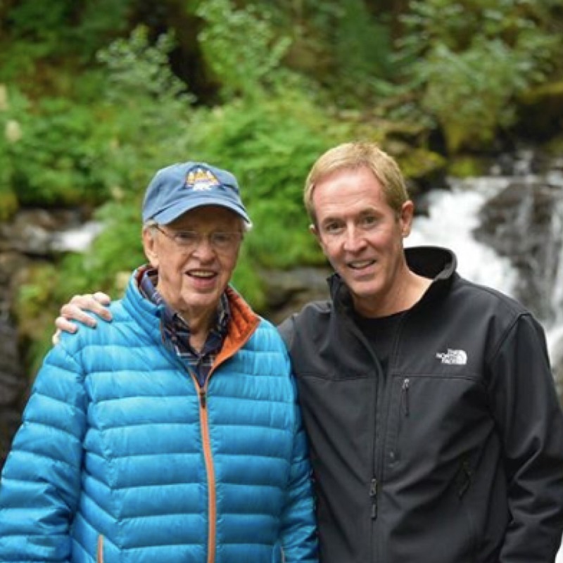Two men outdoors near a waterfall with lush green forest background. Older man wearing blue baseball cap, glasses, and bright blue down jacket; younger man in black North Face jacket. Both are smiling with arms around each other's shoulders.