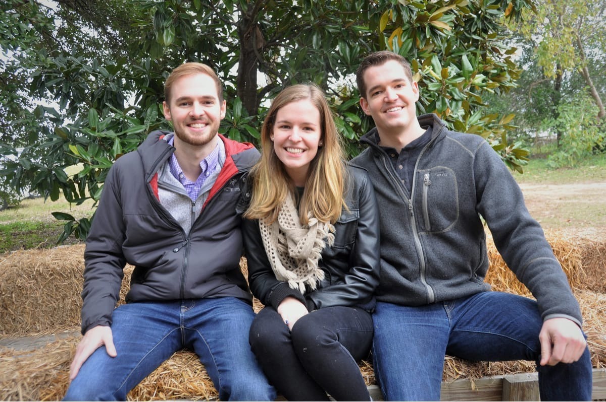 Three young adults sitting on hay bales outdoors, smiling at the camera. Two men wearing jackets (one gray, one dark) flank a woman in the middle wearing a cream-colored scarf. Green trees and foliage visible in the background.