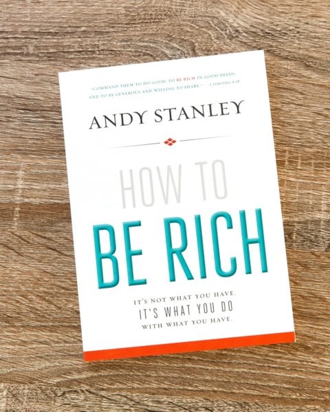 Book cover of "How to Be Rich" by Andy Stanley on wooden surface, with white background and teal title text, subtitle "It's Not What You Have, It's What You Do with What You Have."