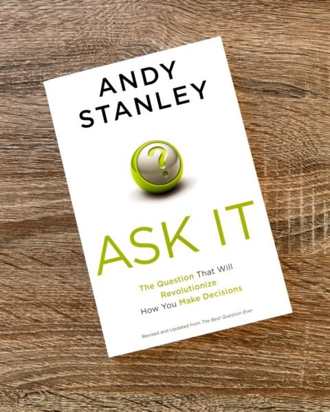 Book cover of "Ask It" by Andy Stanley on wooden surface, featuring white background with green sphere graphic and subtitle "The Question That Will Revolutionize How You Make Decisions.