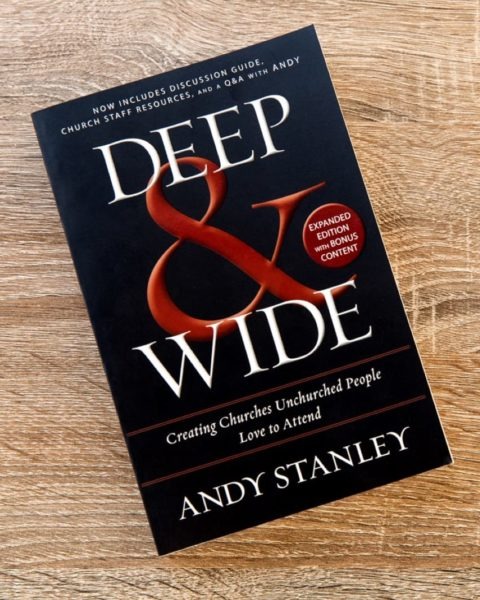 Book cover of "Deep &amp; Wide" by Andy Stanley on wooden surface, featuring black background with red ampersand and subtitle "Creating Churches Unchurched People Love to Attend." Includes "Expanded Edition with Additional Content" badge.