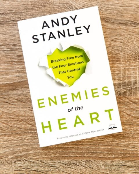 Book cover of "Enemies of the Heart" by Andy Stanley on wooden surface, white background with torn paper effect revealing yellow heart shape, subtitle "Breaking Free from the Four Emotions That Control You."