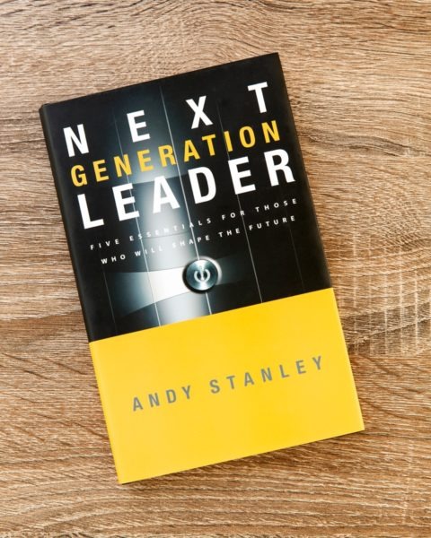 Book cover of "Next Generation Leader" by Andy Stanley on wooden surface, featuring black and yellow design with metallic sphere graphic and subtitle "Five Essentials for Those Who Will Shape the Future."