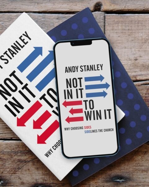 Books and smartphone displaying "Not In It to Win It" by Andy Stanley, with red and blue arrow graphics and subtitle "Why Choosing Sides Sidelines the Church."