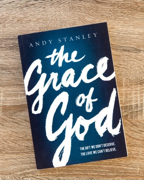 Book cover of "The Grace of God" by Andy Stanley on wooden surface, dark blue background with white handwritten-style title and subtitle "The Gift We Don't Deserve, The Love We Can't Believe."