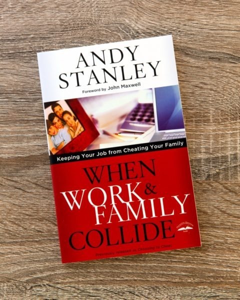 Book cover of "When Work &amp; Family Collide" by Andy Stanley on wooden surface, featuring red and white design with family and computer imagery, subtitle "Keeping Your Job from Cheating Your Family," with foreword by John Maxwell.